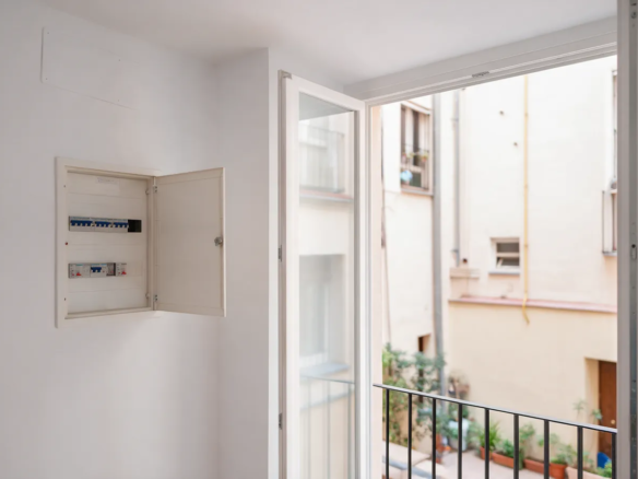 Salón luminoso reformado con ventana a un patio y un cuadro eléctrico abierto en la pared